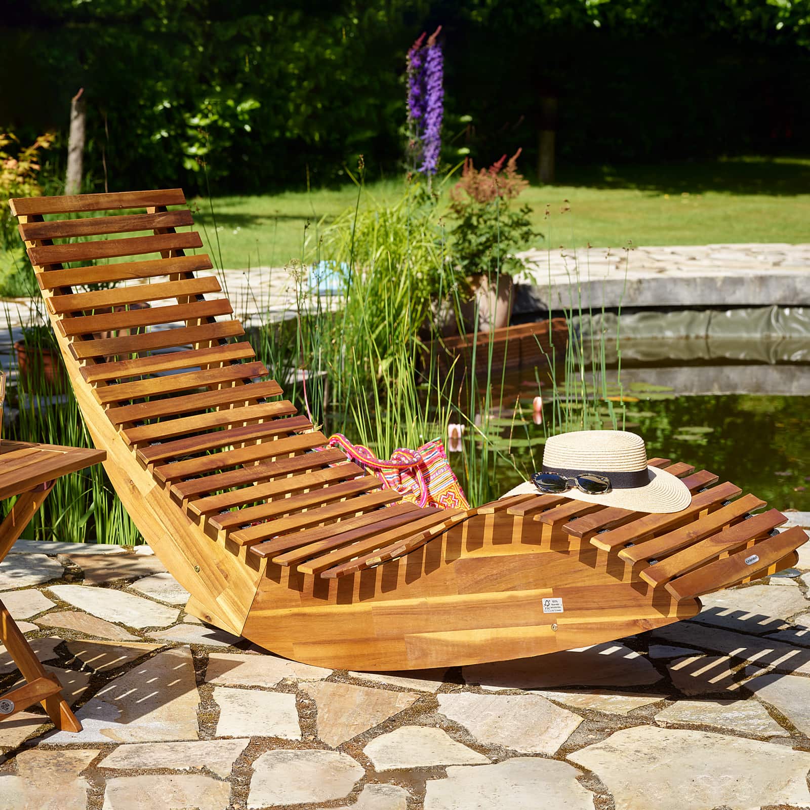 Gyngende liggestol i akacietræ på fast terrasse ved sø, lifestyle billede af fast liggestol-model til varm sommerdag