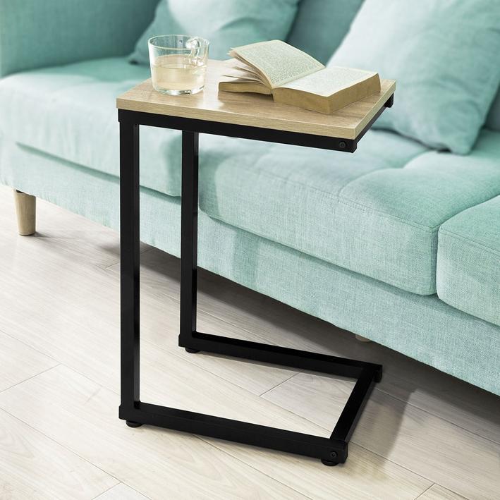 Lille sidebord i lyst træ og sort metal skubbet ind under en mintgrøn sofa med et glas vand og en åben bog ovenpå