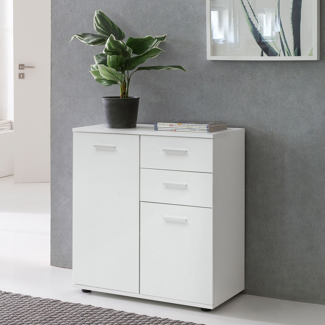 Hvid minimalistisk kommode med låge og skuffer placeret ved grå murvæg med plante og indrammet kunst