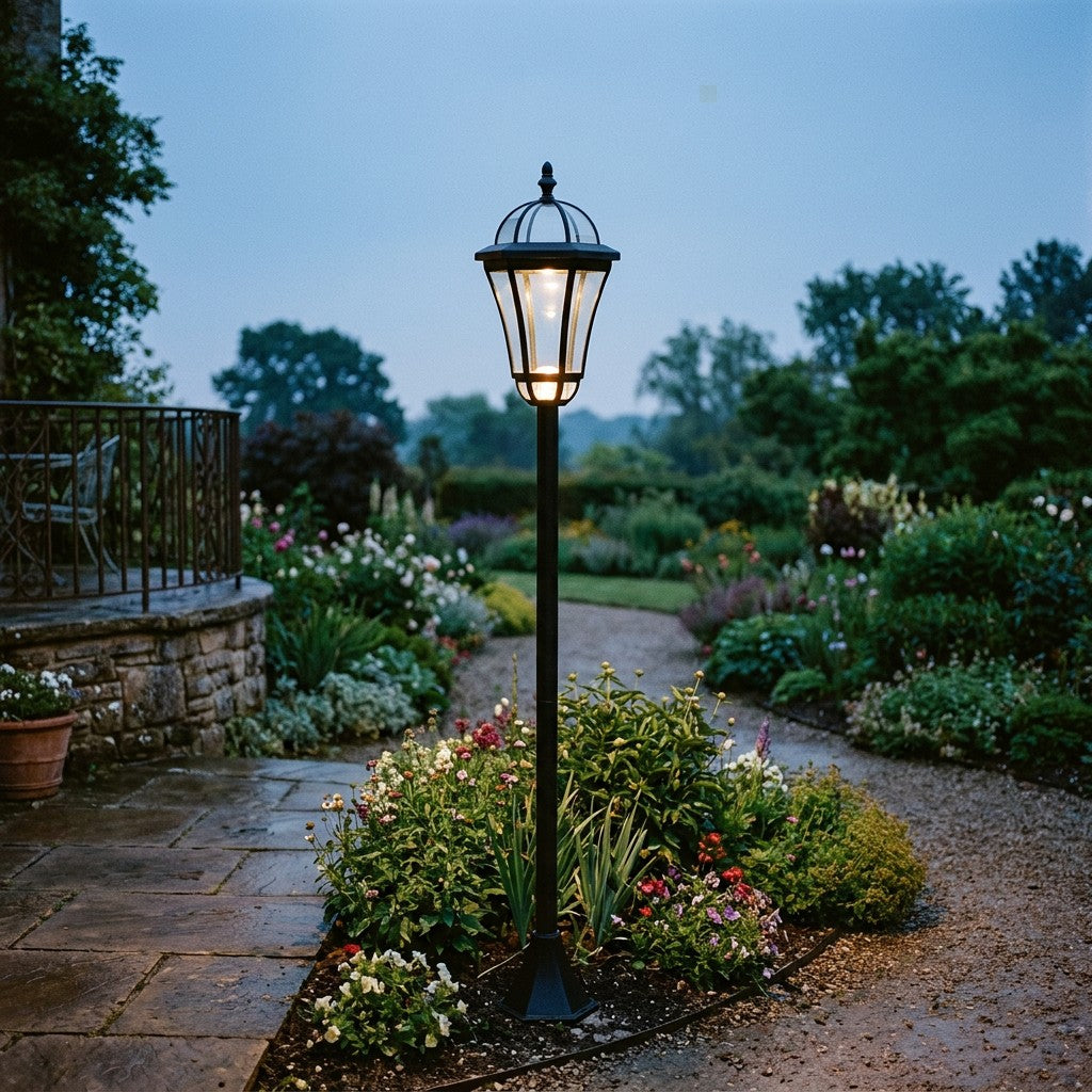 Sort solcellelampe på høj stander med glas-lampehoved, der lyser varmt i et blomsterbed ved en stenterrasse i skumringen